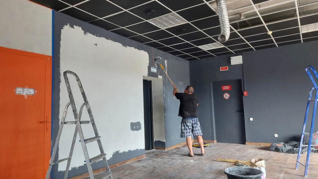 A man is using a roller to paint a wall dark gray in a commercial room. Ladders and painting supplies are arranged around as renovations progress in the space.